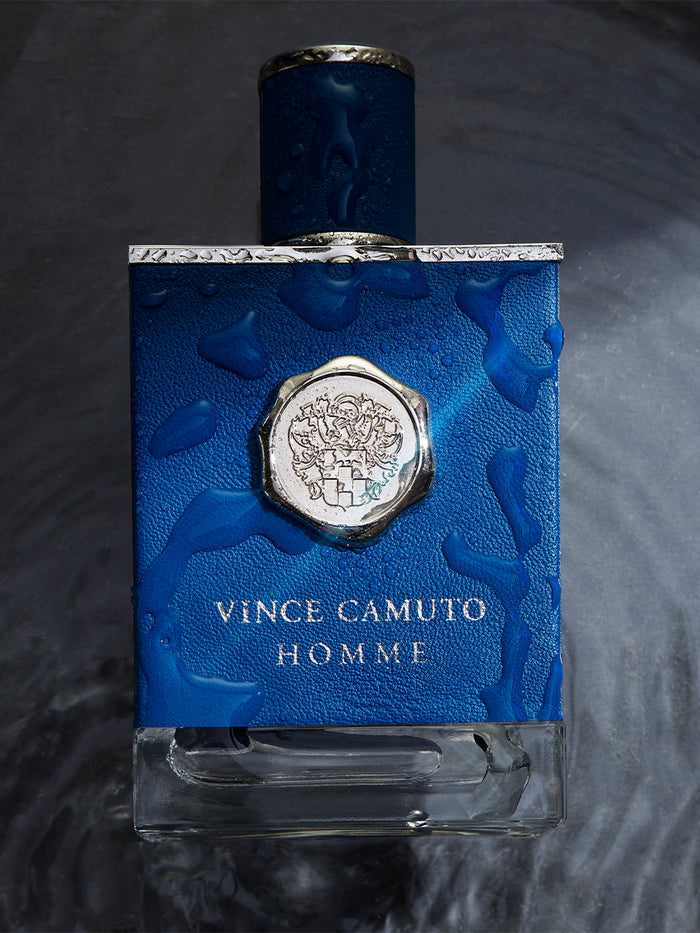 A blue bottle of Vince Camuto Men’s Homme 3.4 oz Eau de Toilette Spray, glistening with water droplets against a wet, dark backdrop, captures the invigorating essence of the Mediterranean and reflects the bold allure of this distinctive men’s fragrance.