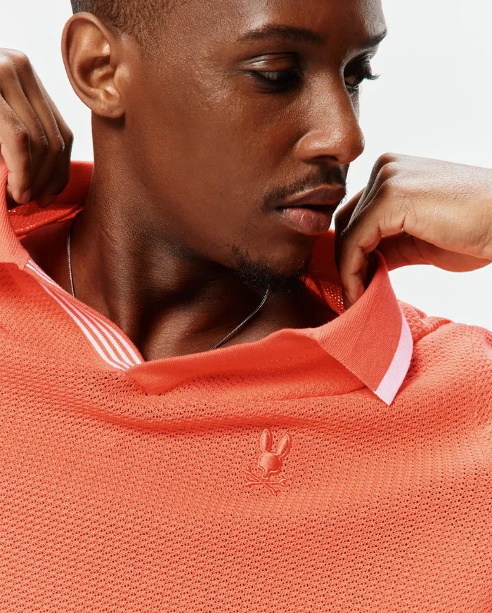 Mens Innis Polo in vibrant coral, featuring a breathable open-knit texture, classic fit, small embroidered Psycho Bunny skull logo on the left chest, and a collar with white stripe accents.