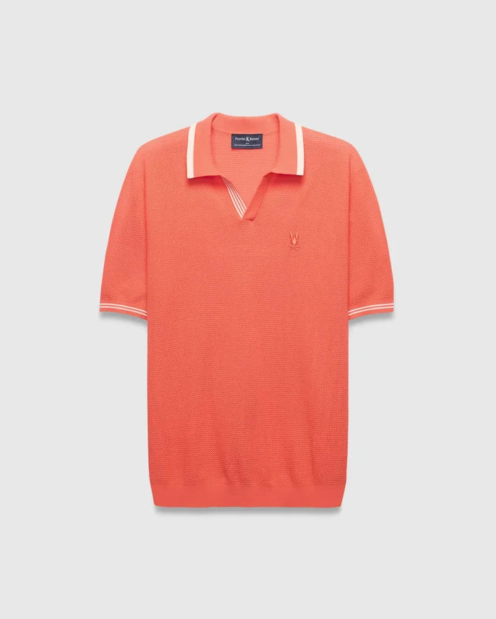 Mens Innis Polo, a vibrant coral knit polo with a V-neck collar and cuffs accented by white stripes, featuring a small embroidered Psycho Bunny skull logo on the left chest. Classic fit.