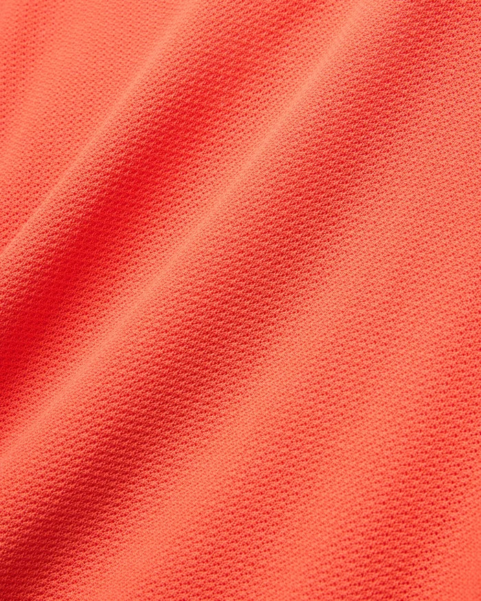 Mens Innis Polo in vibrant coral orange pique knit fabric, showcasing its signature textured, breathable weave with subtle diagonal folds, ideal for premium Psycho Bunny refined streetwear.