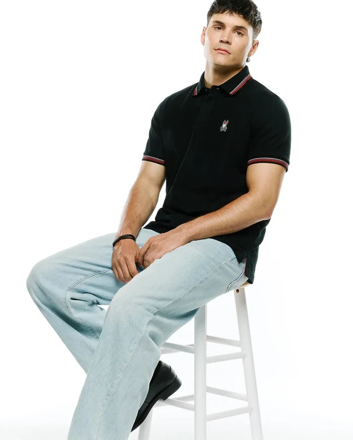 Mens Jaydon Pique Polo in black, featuring a classic fit, textured pique fabric, red and white striped collar and cuffs, and a small embroidered Psycho Bunny skull logo on the left chest.