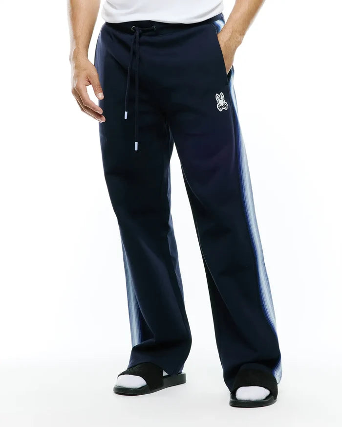 Mens Jaydon Track Pant in navy, featuring a relaxed fit, smooth knit fabric, drawstring waist, and a small embroidered white Psycho Bunny skull logo on the left thigh, with light blue gradient vertical stripes on the sides.