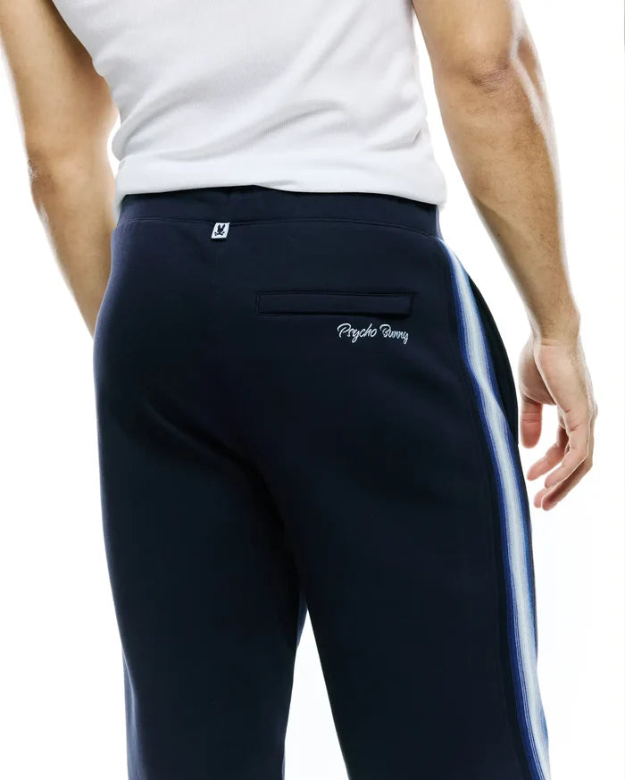 Mens Jaydon Track Pant, navy, relaxed fit jersey, featuring blue/white side stripes, small bunny skull logo tag on back waistband, and embroidered Psycho Bunny wordmark on back pocket.