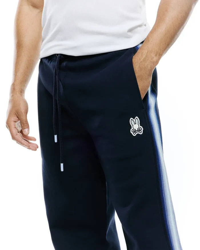 Mens Jaydon Track Pant in navy, featuring a relaxed fit, smooth knit fabric, drawstring waist, and a small embroidered Psycho Bunny skull logo on the left thigh, with a blue and white striped side panel.