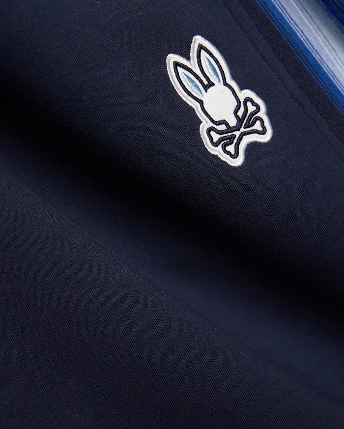 Navy blue Mens Jaydon Track Pant in smooth knit, featuring a prominent embroidered white and light blue Psycho Bunny skull logo and a multi-tonal blue vertical side stripe.