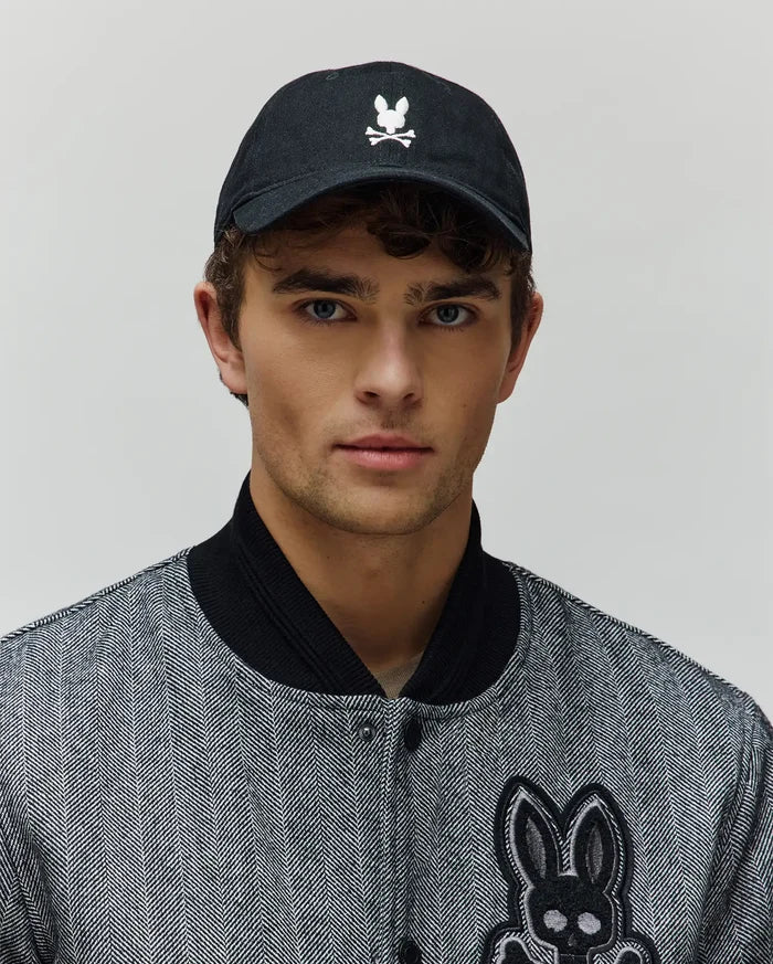 Jimmy Baseball Cap in black with white embroidered Psycho Bunny skull logo, paired with a black and white herringbone bomber jacket featuring a large embroidered Psycho Bunny patch.