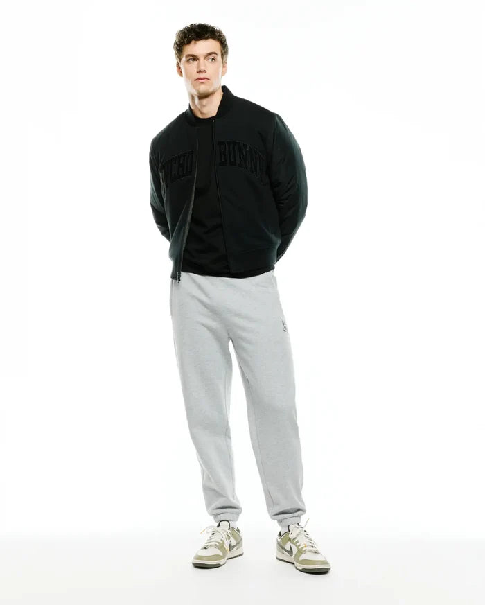 Mens Joaquin Mix Media Reversible Bomber in black, featuring large tonal Psycho Bunny lettering across the chest, paired with light grey relaxed fit french terry sweatpants with a small embroidered bunny skull logo on the left thigh.