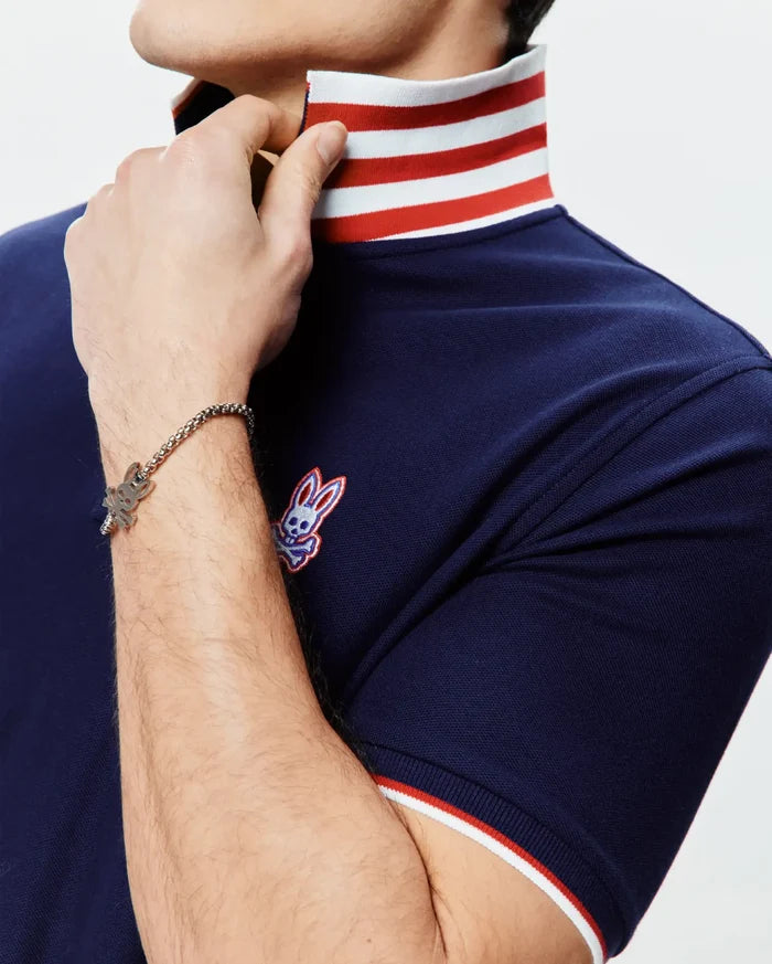 Mens John Pique Polo, a navy blue pique polo with a classic fit, featuring a white and red striped collar and sleeve cuffs, and an embroidered Psycho Bunny skull logo on the left chest.