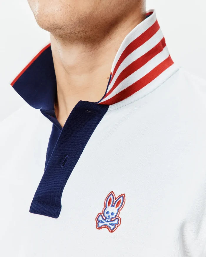 Mens John Pique Polo, a classic fit light blue pique polo with a navy placket, a bold red and white striped collar, and an embroidered Psycho Bunny skull logo on the left chest.