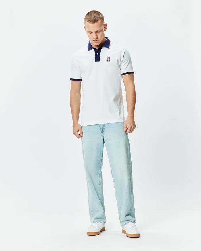 Mens John Pique Polo, a classic fit white pique polo with a navy collar, two-button placket, red and navy striped sleeve cuffs, and a small embroidered Psycho Bunny skull logo on the left chest.