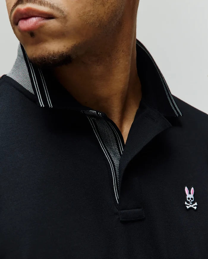 Mens Kenyon Lightweight Pique Polo in black, featuring a classic fit, textured pique fabric, and a small embroidered Psycho Bunny skull logo with pink ears on the left chest. The collar has a subtle houndstooth pattern and white stripe accents.