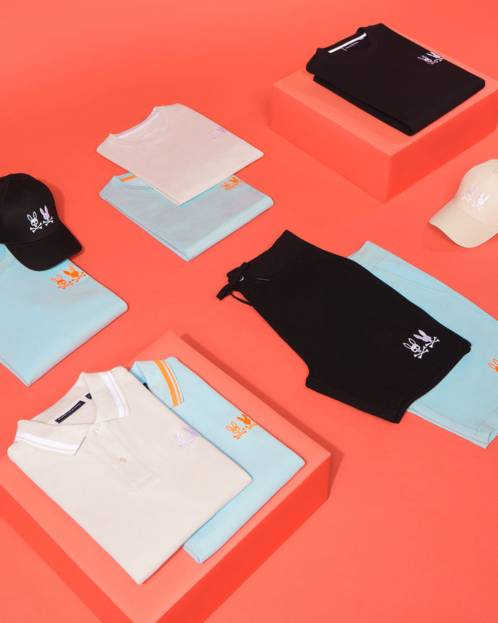 An arrangement of clothing and accessories on a light red background. Items include light blue and black shorts, white and black t-shirts, light blue polo shirts with orange and white graphics, and Psycho Bunny MENS KINGWOOD EMBROIDERED BASEBALL CAP - B6A916Y1HT with adjustable backstraps. The majority feature an embroidered rabbit logo.