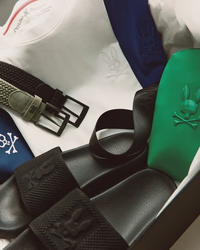 White jersey t-shirt, navy garment, green pouch, and Black Mens Knit Slides with textured knit strap, all featuring the iconic Psycho Bunny skull logo. Olive and black woven belts.
