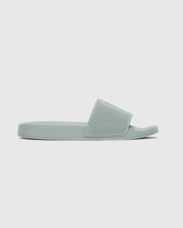 The Psycho Bunny MENS KNIT SLIDES - B6A474F200 feature a light green knit upper, textured wide strap, flat sole, and ergonomically molded footbed, shown in side profile against a plain light background.