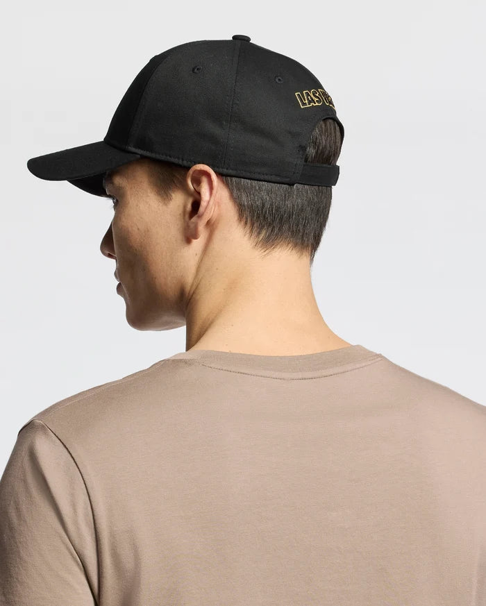 Seen from behind, a person wears the MENS LAS VEGAS BASEBALL CAP - MO1500011 by Psycho Bunny, featuring "LAS VEGAS" in yellow direct embroidery on black. Their short dark hair contrasts with a beige 100% cotton T-shirt against a plain, light background.