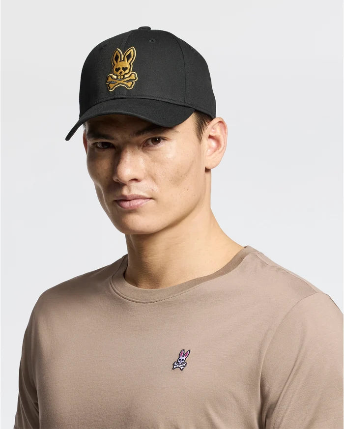 A person wears a beige 100% cotton t-shirt with a small rabbit logo, paired with the Psycho Bunny MENS LAS VEGAS BASEBALL CAP in black, featuring a direct embroidery yellow skull and crossbones rabbit logo. The background is plain white.