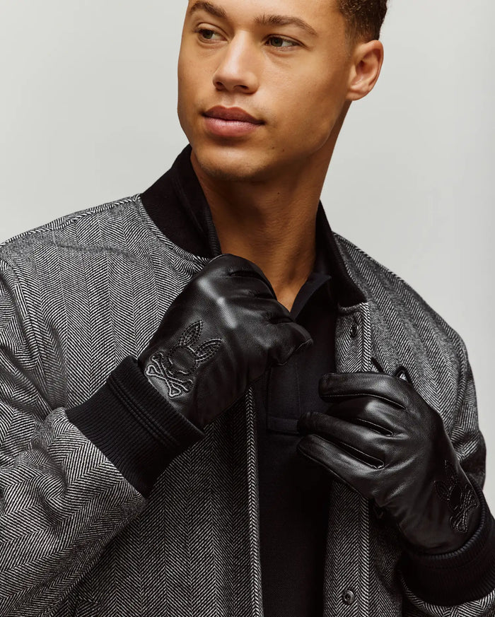 Black and white herringbone bomber jacket with black ribbed cuffs and collar, worn over a black polo shirt, paired with black Mens Leather Gloves featuring an embossed Psycho Bunny skull logo.