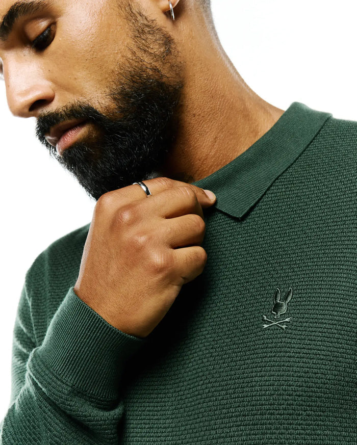 Mens Leroy Long Sleeve Knit Polo in deep forest green, featuring a textured knit fabric, ribbed collar and cuffs, and a small, tonal embroidered Psycho Bunny skull logo on the left chest.