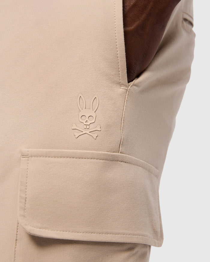 Close-up of a pocket on the MENS LLOYD CARGO JOGGER - B6P159B200 by Psycho Bunny, showcasing a stitched logo of a bunny head with crossbones underneath. The smooth fabric pocket has a flap secured by snap fastening, and a hand is casually resting in the side pocket, highlighting the refined embroidery.