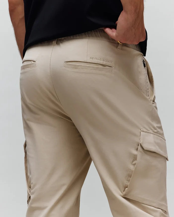 Mens Luka Cargo Pant in light beige, featuring a relaxed fit, smooth fabric, large side cargo pockets, rear welt pockets, and a subtle embroidered Psycho Bunny text logo above the right back pocket.
