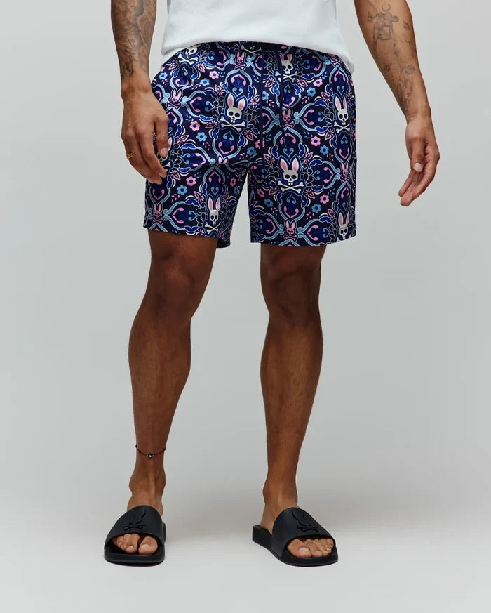 Mens Luke Swim Trunk, a classic fit, navy blue swim trunk with an all-over ornate floral and paisley pattern in pink, light blue, purple, and white, featuring integrated Psycho Bunny skull logos.