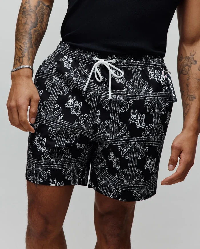 Mens Luke Swim Trunk, black with an all-over white paisley bandana print featuring the iconic Psycho Bunny skull logo, white drawstring, and a branded Psycho Bunny tag.
