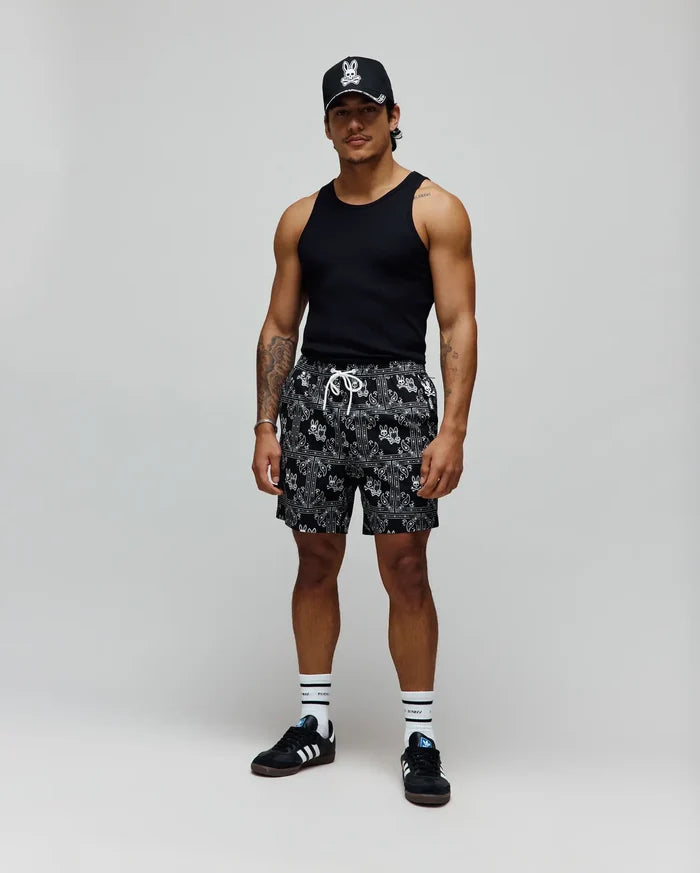Mens Luke Swim Trunk in black with white bandana print featuring repeated small Psycho Bunny skull logos and white drawstring. Worn with a black tank top and black cap with large white Psycho Bunny logo.