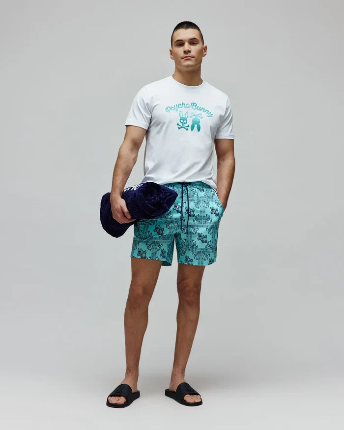 White jersey t-shirt with a large teal gradient Psycho Bunny graphic on the chest, paired with Mens Luke Swim Trunk in aqua blue featuring an all-over dark blue bandana print with repeated bunny skull logos and a black drawstring.