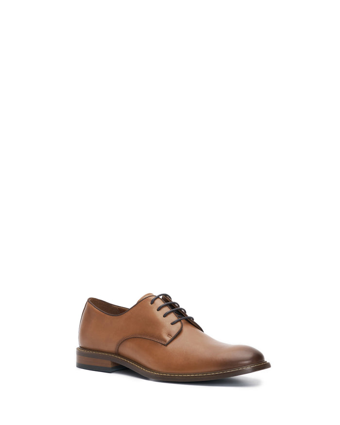 The Vince Camuto Men’s Lyre Derby is a tan leather dress shoe for men, featuring black laces and a low heel. Shown at a slight angle on a white background, this elegant design embodies timeless style.