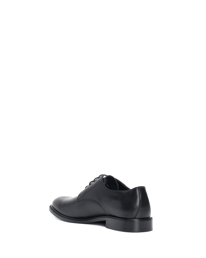 The Vince Camuto Men’s Lyre Derby shoe, crafted from dark leather, is displayed from the back and side on a white background. This classic men’s black dress shoe features laces, a low heel, and timeless derby styling.