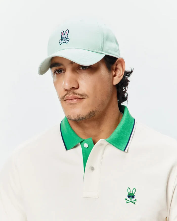 Off-white pique polo with bright green collar, navy stripe, and embroidered Psycho Bunny skull logo with sunglasses on chest. Light mint Mens Classic Baseball Cap with embroidered Psycho Bunny logo.