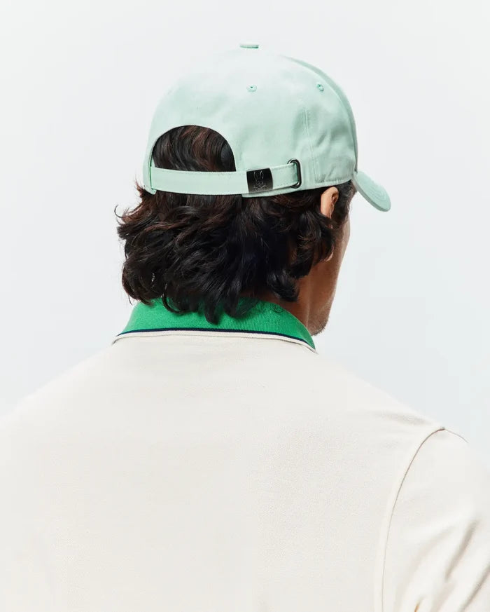 Mint Green Mens Classic Baseball Cap with a Psycho Bunny skull logo on the metal buckle, paired with a cream pique polo shirt featuring a green ribbed collar and navy stripe.
