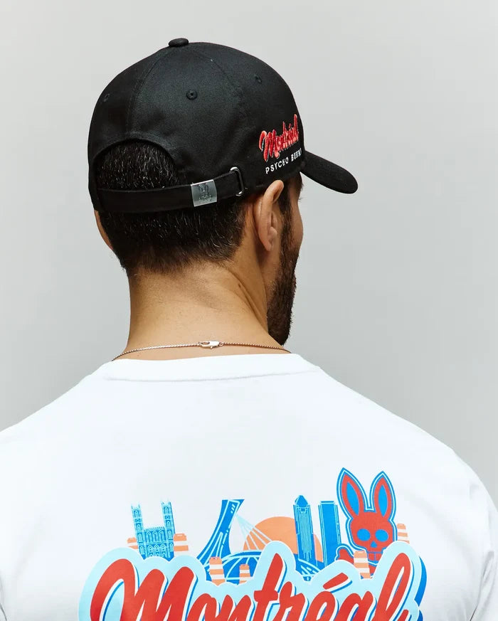 A person wears a black Psycho Bunny MENS MONTREAL BASEBALL CAP (MO1500048) and a white T-shirt with colorful Montreal graphics on the back, standing with their back to the camera against a plain light background.