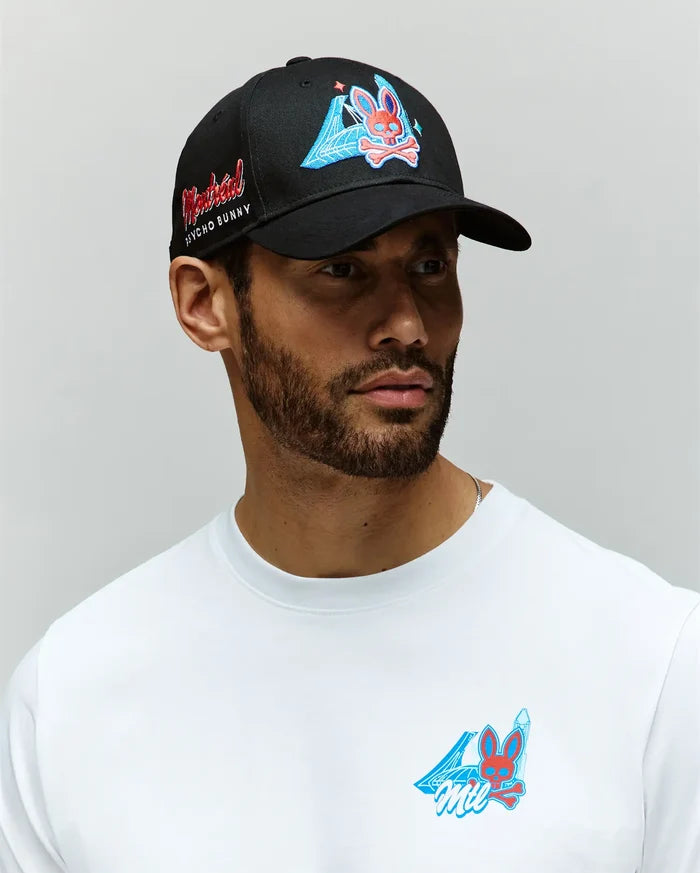 A bearded man wears a white shirt and a Psycho Bunny MENS MONTREAL BASEBALL CAP (MO1500048), both with colorful cartoon bunny graphics and blue designs. The black 100% cotton cap features an embroidered Bunny and red side text. The background is plain gray.