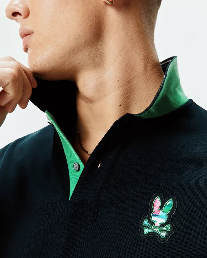 Mens Nate Pique Polo, a dark navy classic fit pique polo featuring a vibrant green contrast collar and a prominent multi-color embroidered Psycho Bunny skull logo on the chest.