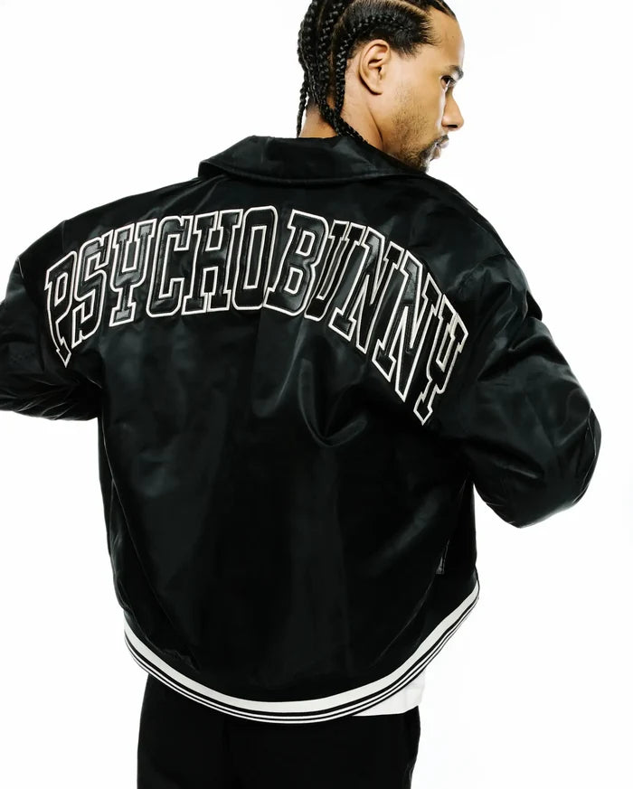 Black Mens Nathaniel Satin Coach Jacket, a relaxed fit, smooth satin jacket with a large white outlined PSYCHO BUNNY text logo across the upper back and a ribbed hem with white double stripes.