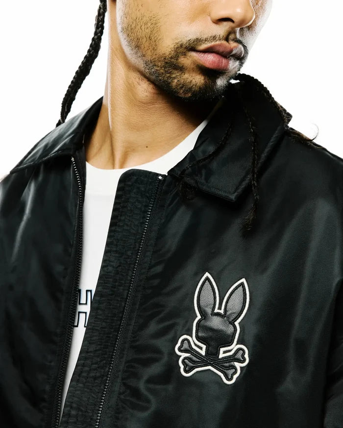 Mens Nathaniel Satin Coach Jacket, black with a subtle sheen, featuring a prominent embroidered Psycho Bunny skull logo on the left chest with white outline and black textured fill, and a full zip.