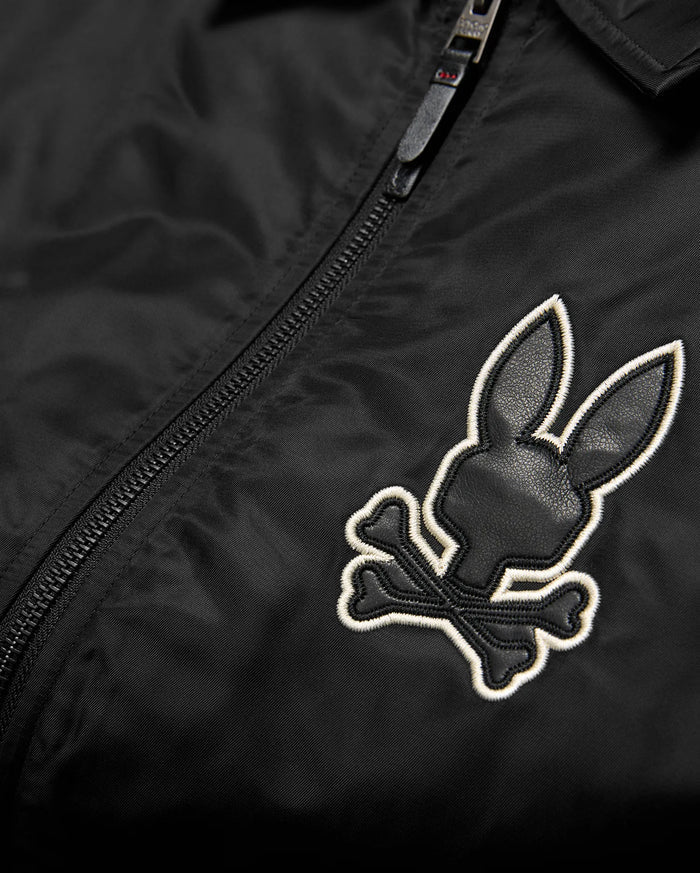 Mens Nathaniel Satin Coach Jacket in black, featuring a prominent embroidered Psycho Bunny skull logo on the chest with black leather-like texture and a white outline, alongside a black zipper.