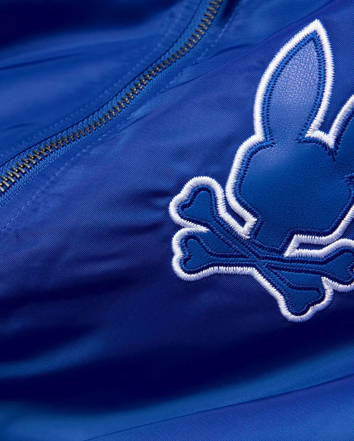 Mens Nathaniel Satin Coach Jacket in royal blue satin, featuring a large, embroidered Psycho Bunny skull logo with a dark blue textured fill and thick white outline, alongside a silver zipper.