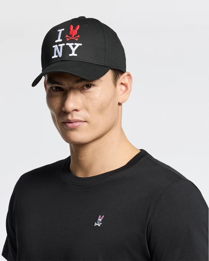 A man wearing a black cotton t-shirt and a Psycho Bunny MENS NEW YORK BASEBALL CAP (MO1500012) with "I ♥️ NY" embroidered in white and red stands against a plain background.