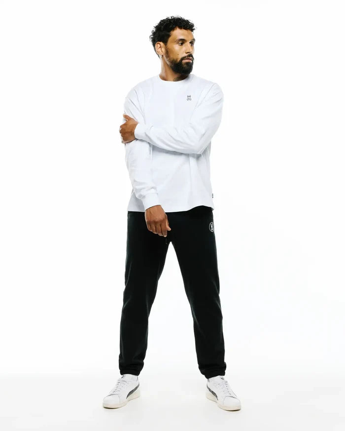 Mens Norman Long Sleeve Oversized Fit Tee in white jersey knit with a small embroidered Psycho Bunny skull logo on the chest, paired with black French terry joggers featuring a white circular bunny logo on the left thigh.