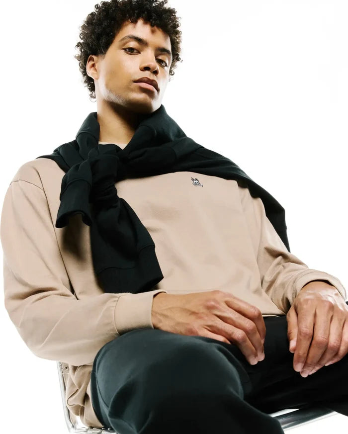 Mens Norman Long Sleeve Relaxed Fit Tee in beige jersey, featuring a small embroidered black Psycho Bunny skull logo on the left chest, paired with black pants and a black sweater tied over the shoulders.