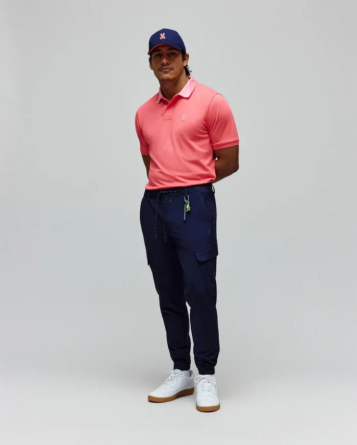 Mens Onyx Sport Polo in coral pique with a lighter pink collar and embroidered chest logo, paired with navy slim-fit cargo joggers featuring a tonal bunny logo and a navy cap with a pink embroidered bunny.