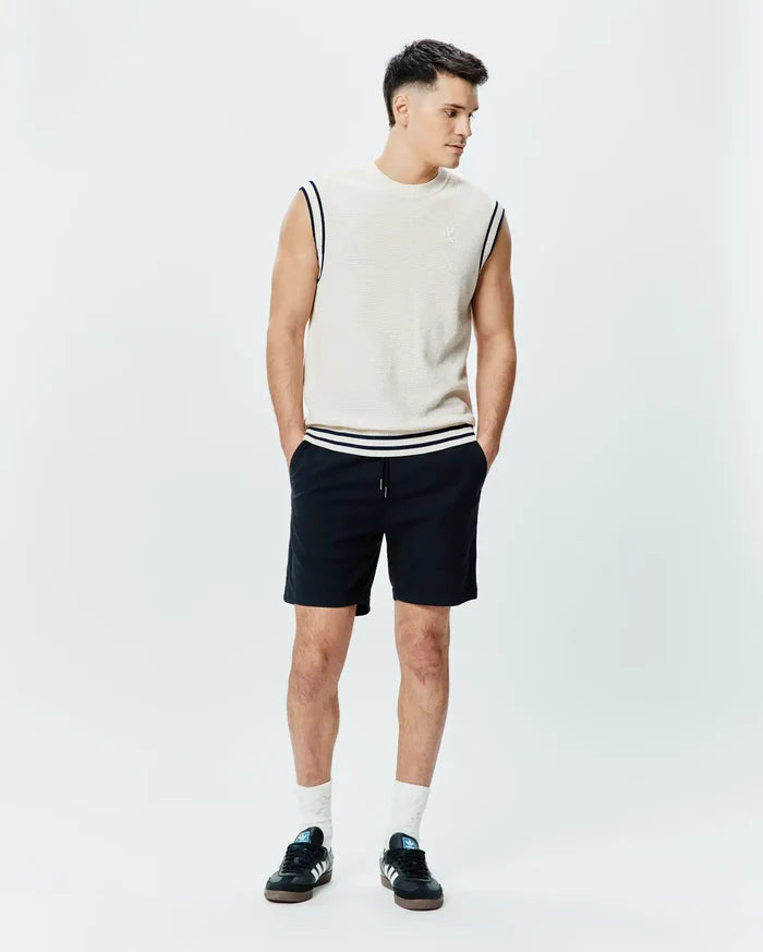 Mens Peter Knit Vest in cream, featuring a classic fit, fine knit texture, navy and white striped armhole and hem trim, and a small embroidered Psycho Bunny skull on the left chest, paired with black drawstring shorts.