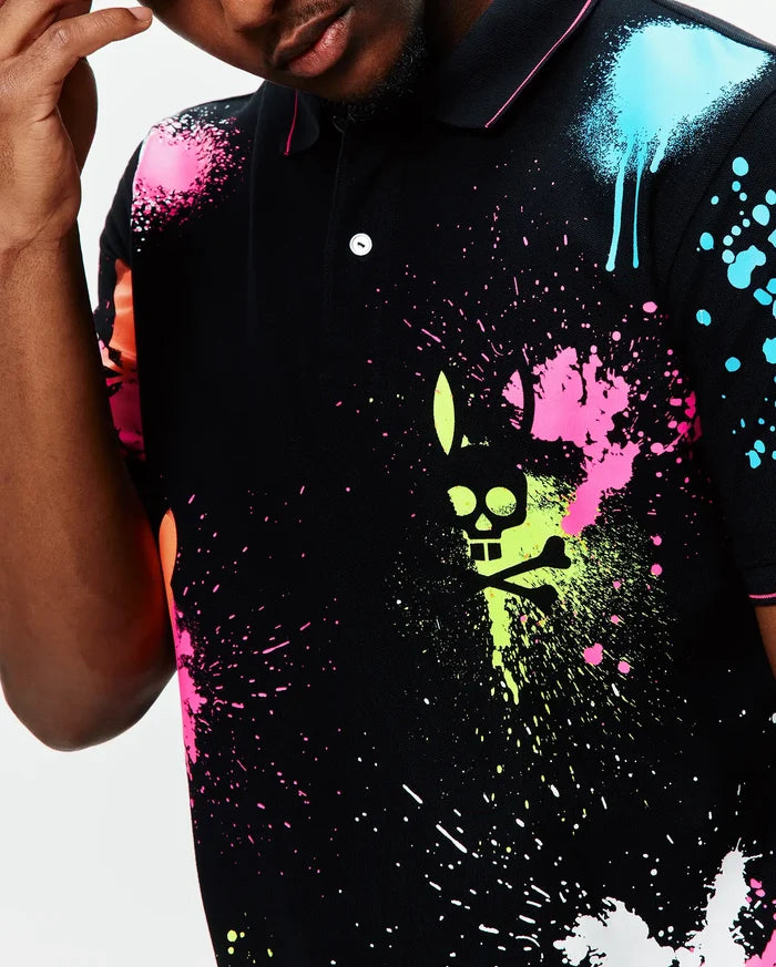 Mens Quintin Aop Pique Polo, a black pique polo with a vibrant all-over neon paint splatter print in pink, lime, blue, and orange, featuring a large central Psycho Bunny skull logo graphic.