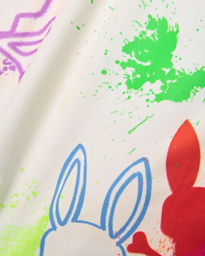 Mens Quintin All Over Print Swim Trunk, cream fabric with vibrant green, purple, blue, and red paint splatter print, featuring repeated Psycho Bunny skull outlines and solid shapes.