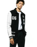 Mens Reilly Knit Coach Jacket, a classic fit black and white varsity-style knit jacket with embroidered 05, Psycho Bunny patches, New York text, vertical sleeve branding, and striped cuffs.