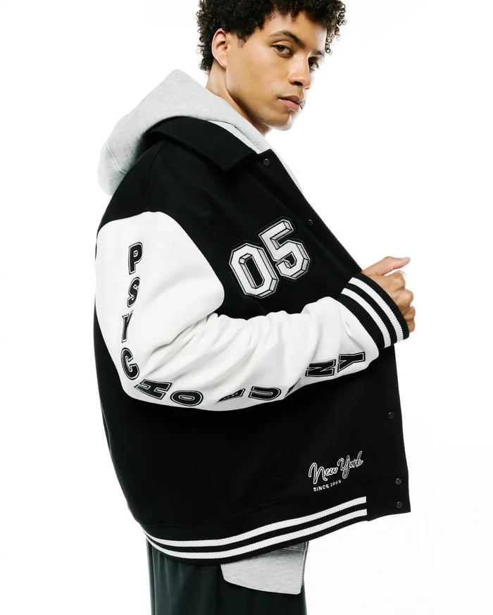 Mens Reilly Knit Coach Jacket, black with white sleeves, large embroidered 05, vertical PSYCHO BUNNY text, New York Since 2005 script, striped ribbed cuffs/hem, layered over a grey hoodie.