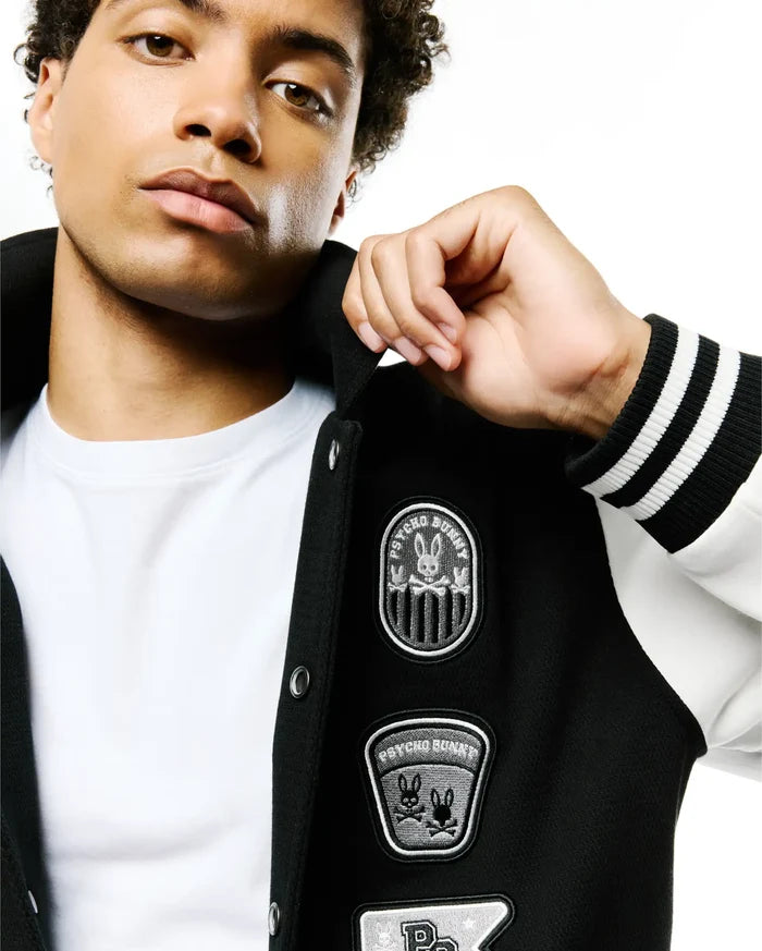 Mens Reilly Knit Coach Jacket, black with white sleeves, features multiple embroidered Psycho Bunny skull patches on the chest, ribbed cuffs with white stripes, and a classic fit.