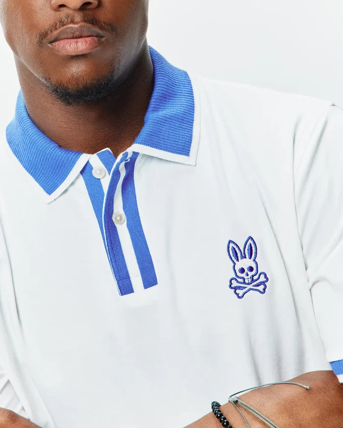 Mens Ridley Pique Polo in white pique, featuring a classic fit, ribbed blue collar, striped blue and white placket, and an embroidered blue Psycho Bunny skull logo on the left chest.